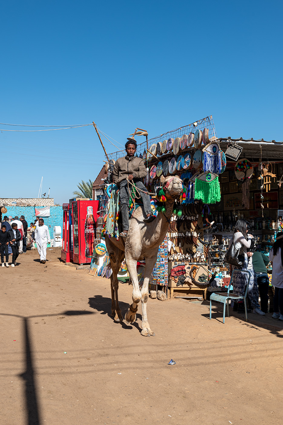 A kid riding a camel in a street with shop vendors